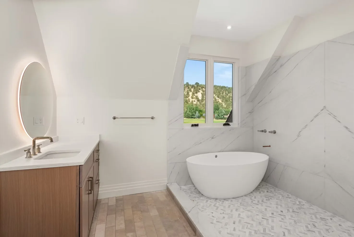 Modern bathroom with marble walls, freestanding tub, wood vanity, and backlit mirror