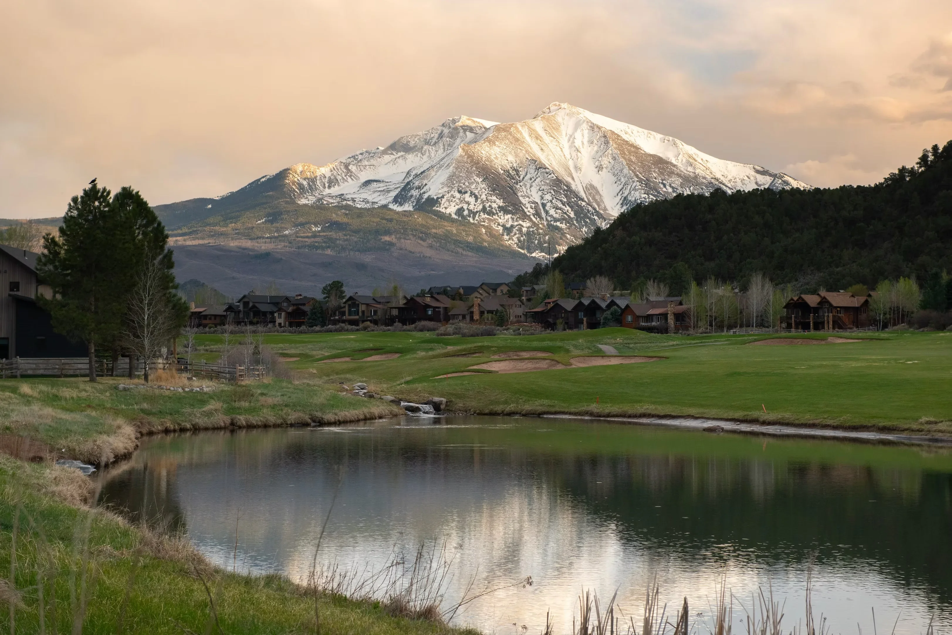 Custom homes in mountain valley with snow-capped peak, green meadow, and stream