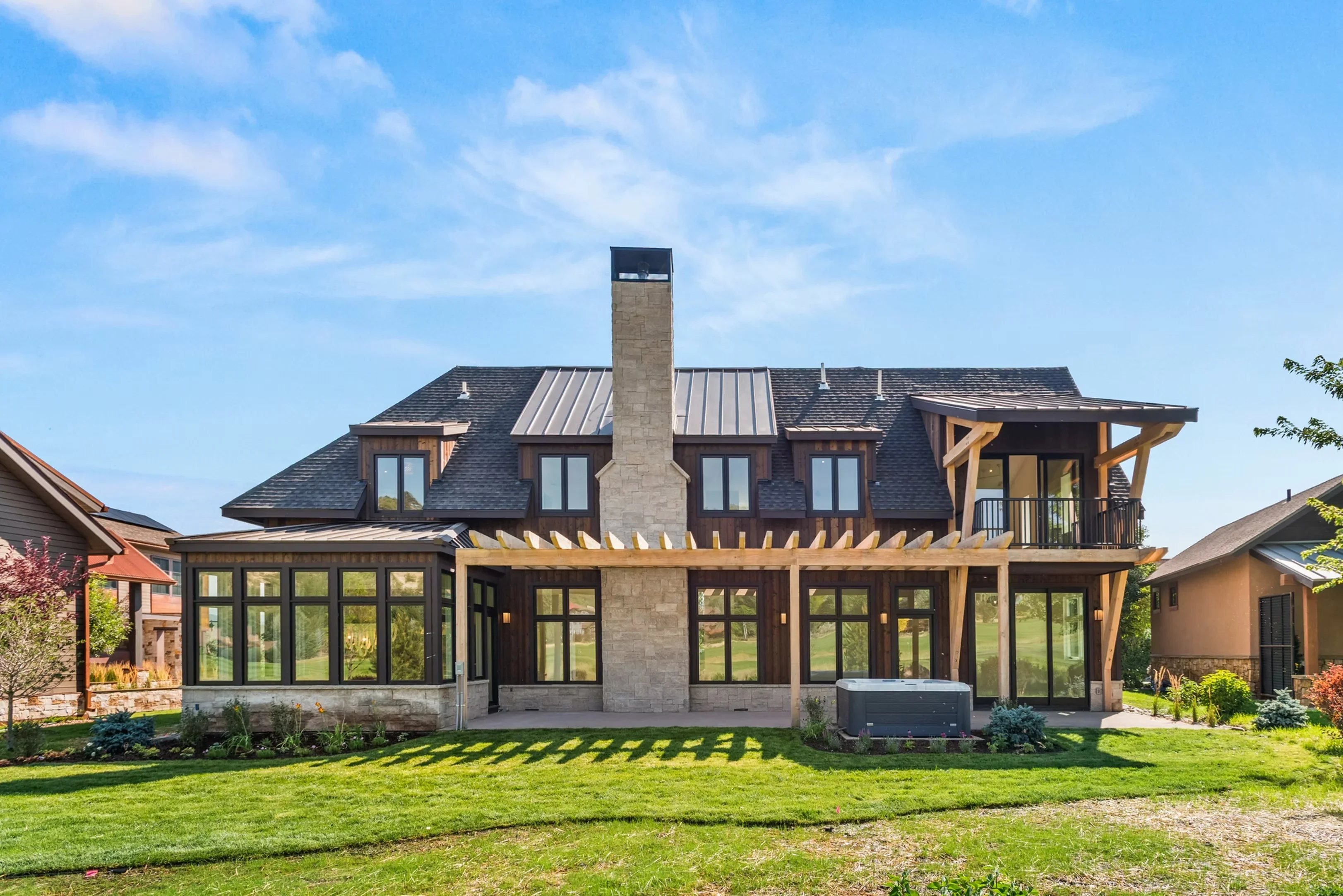 Modern farmhouse with stone chimney, metal roof, black-framed windows, timber pergola, and rear patio