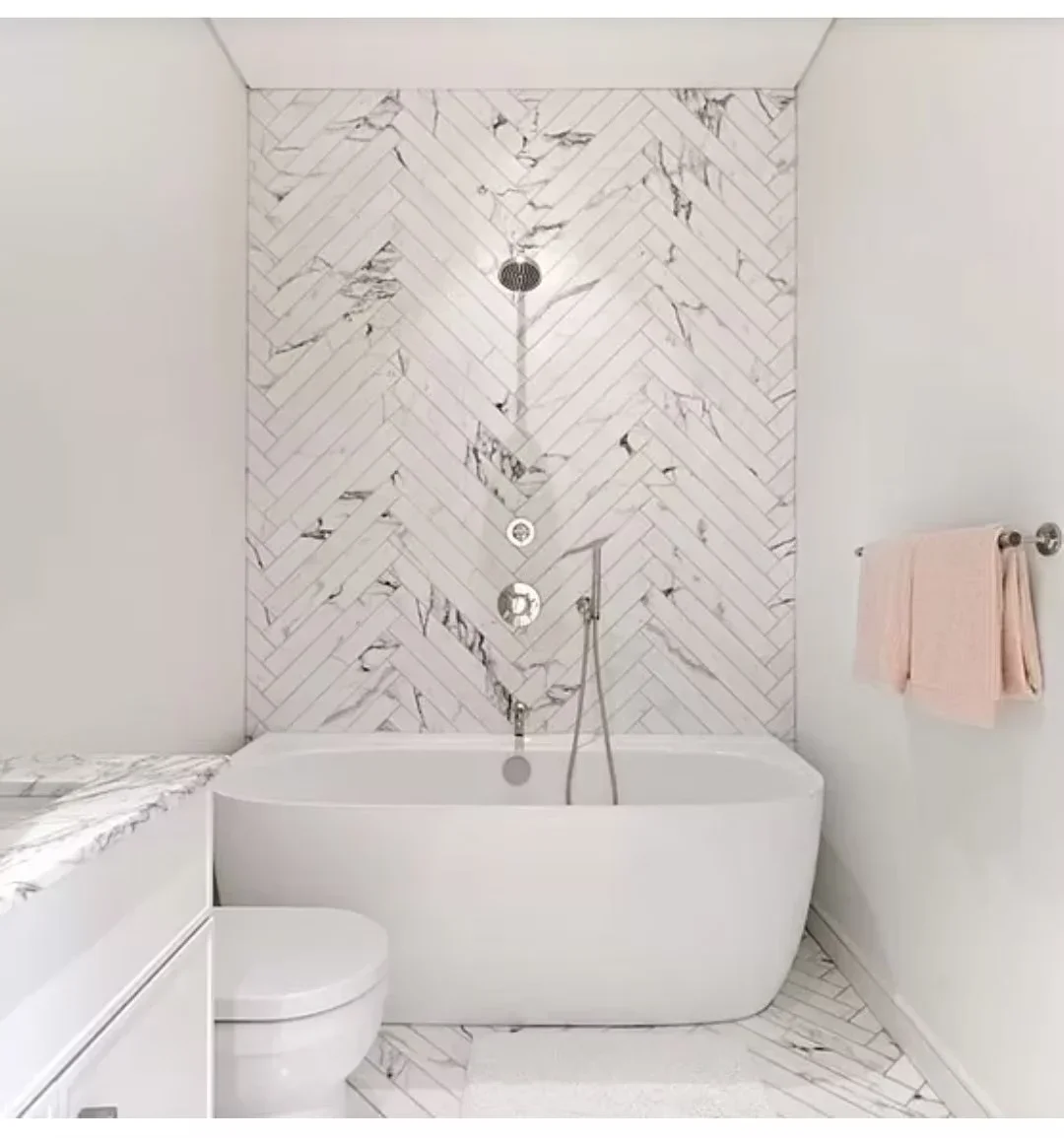 Freestanding white soaking tub with marble herringbone tile wall, chrome rainfall shower, and marble flooring