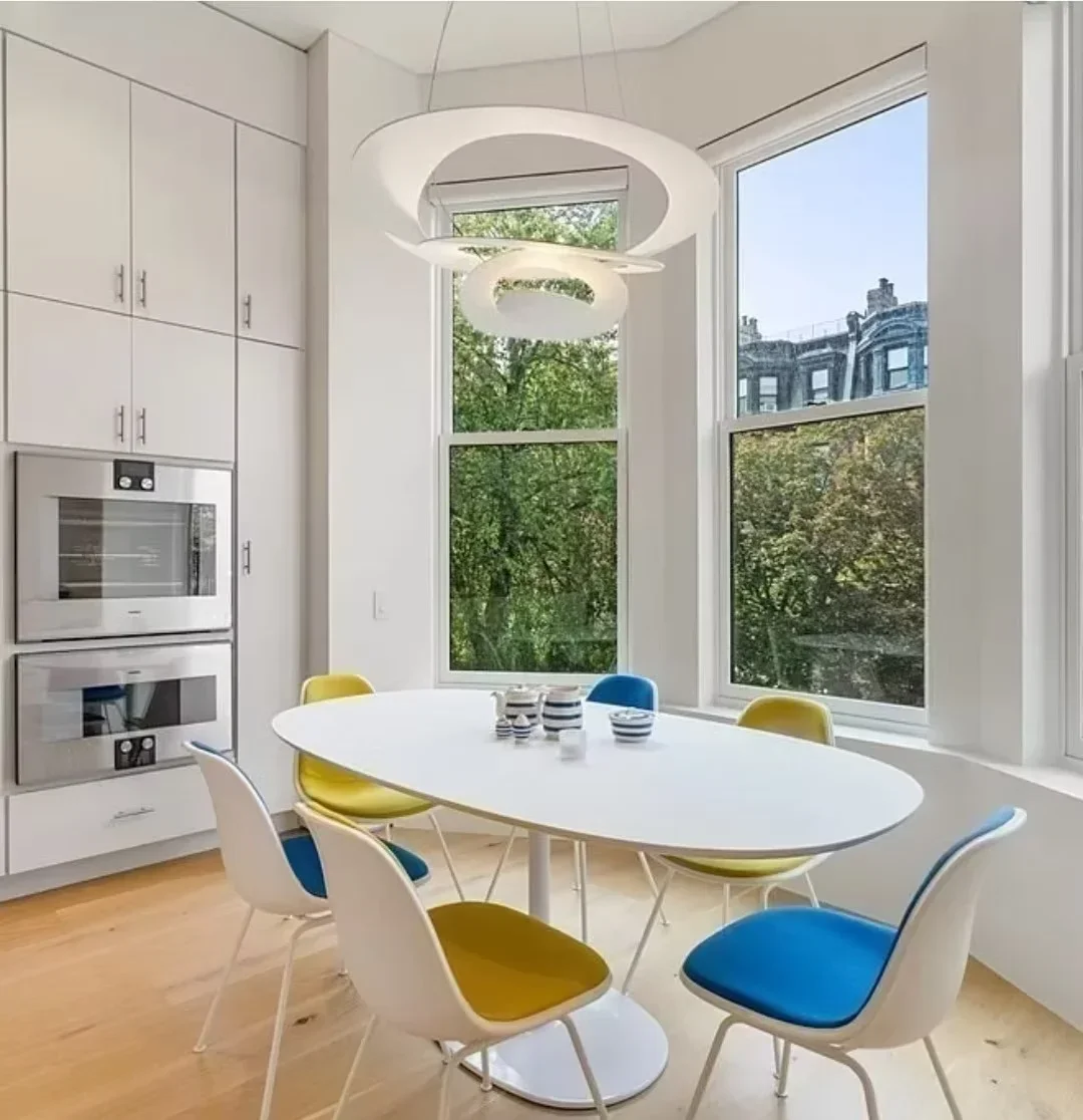 Modern kitchen with white cabinetry, stainless steel ovens, oval dining table, and large windows